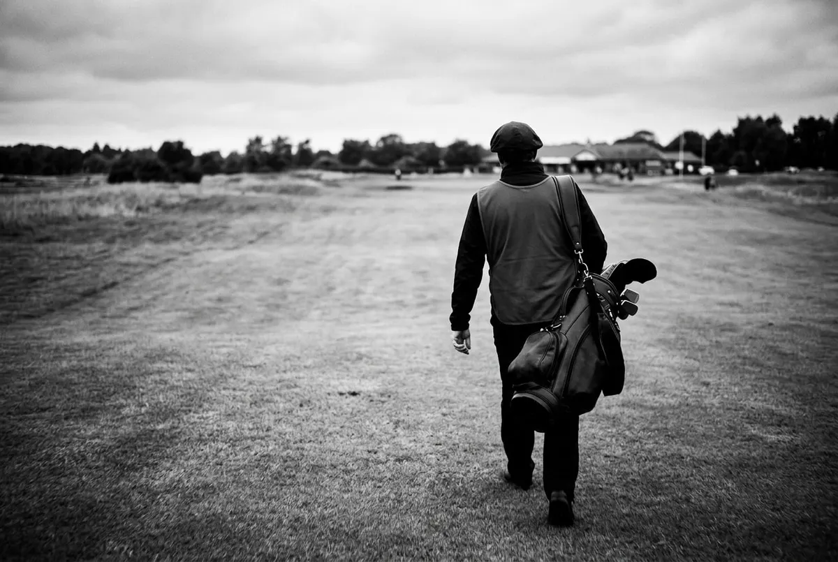 Een caddie loopt alleen met zijn tas over de fairway, van achteren gefotografeerd onder een bewolkte lucht.