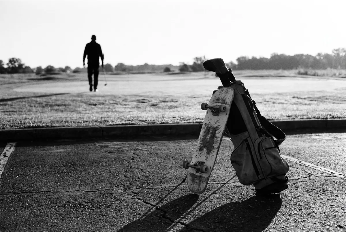Een skateboard leunt tegen een golftas op de grens tussen asfalt en een golfbaan, met een silhouet dat wegloopt richting de green.