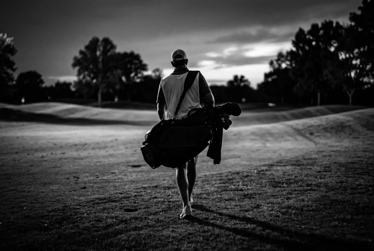 Een caddie met een golftas over zijn schouder loopt over de fairway, gezien van achteren.