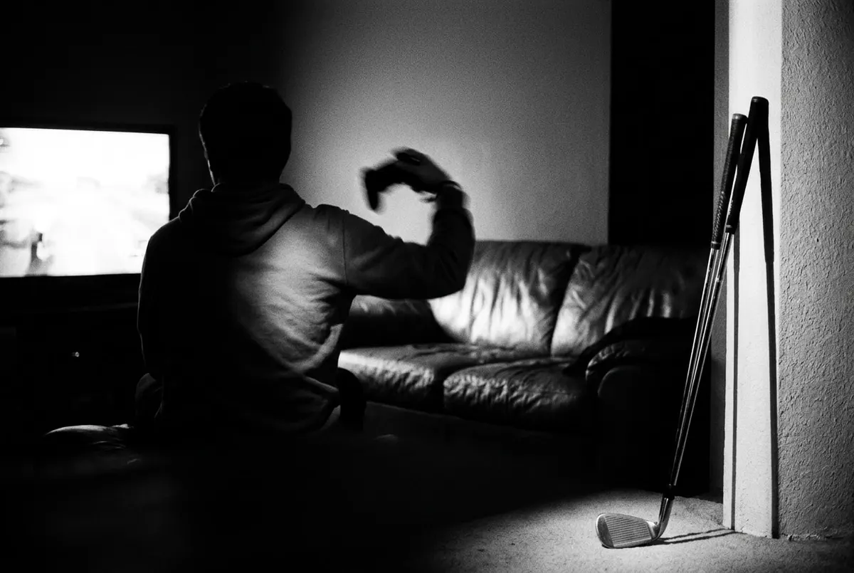 Silhouet van iemand met een controller voor een gloeiend scherm, met op de voorgrond een echte golfclub tegen de muur.
