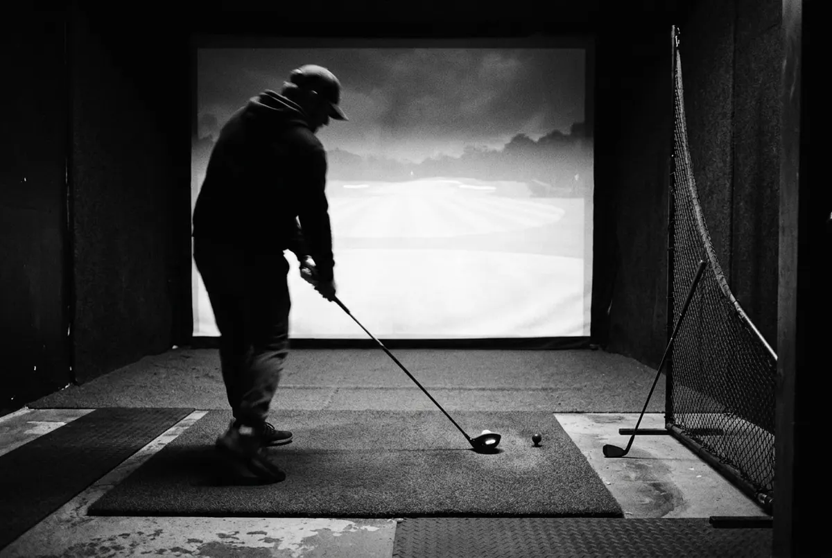 Zwart-witfoto van een indoor golf simulator-bay met een oplichtend projectiescherm en een silhouet in swing.