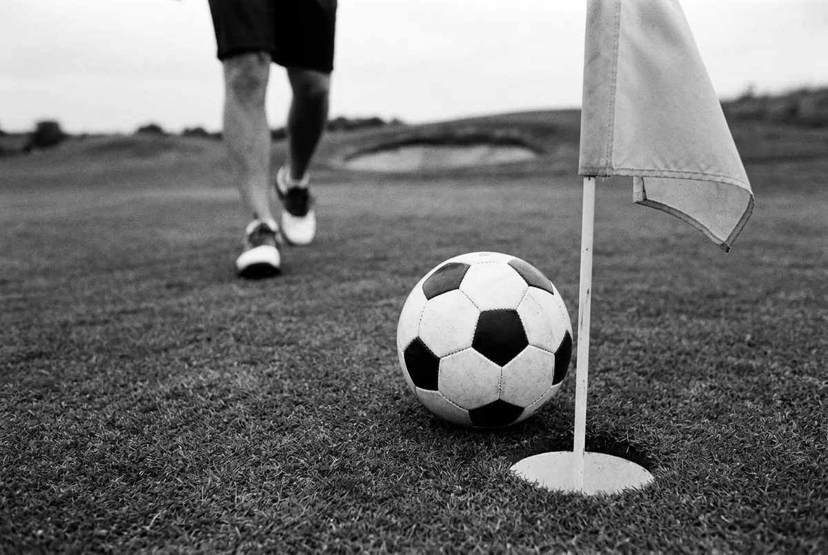 Een voetbal ligt naast de hole op een voetgolfbaan, met een speler in sportschoenen op de achtergrond.