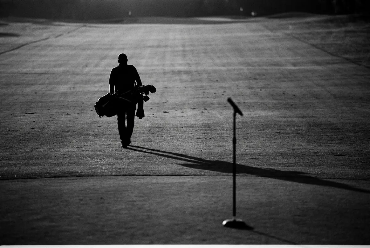 Silhouet van een caddie met golftas op een fairway, met een microfoonstandaard op de plek van de vlag