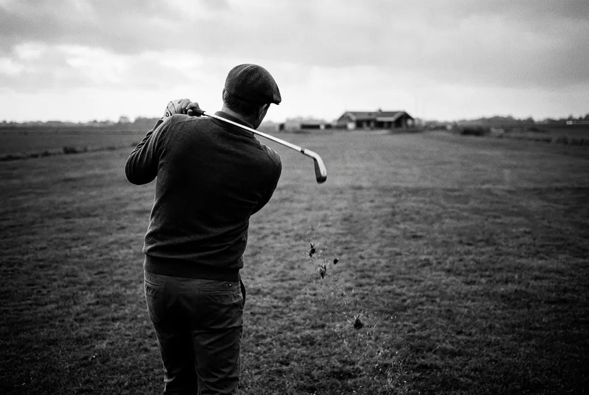 Golfer van achteren slaat met een ijzer-7 op een rustige, bewolkte Nederlandse golfbaan in de herfst