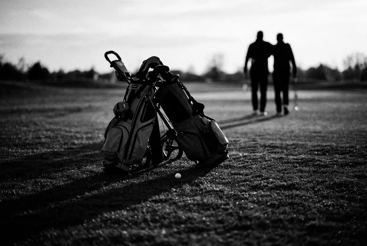 Twee golftassen tegen één trolley met daartussen één golfbal, silhouetten van twee spelers op de achtergrond