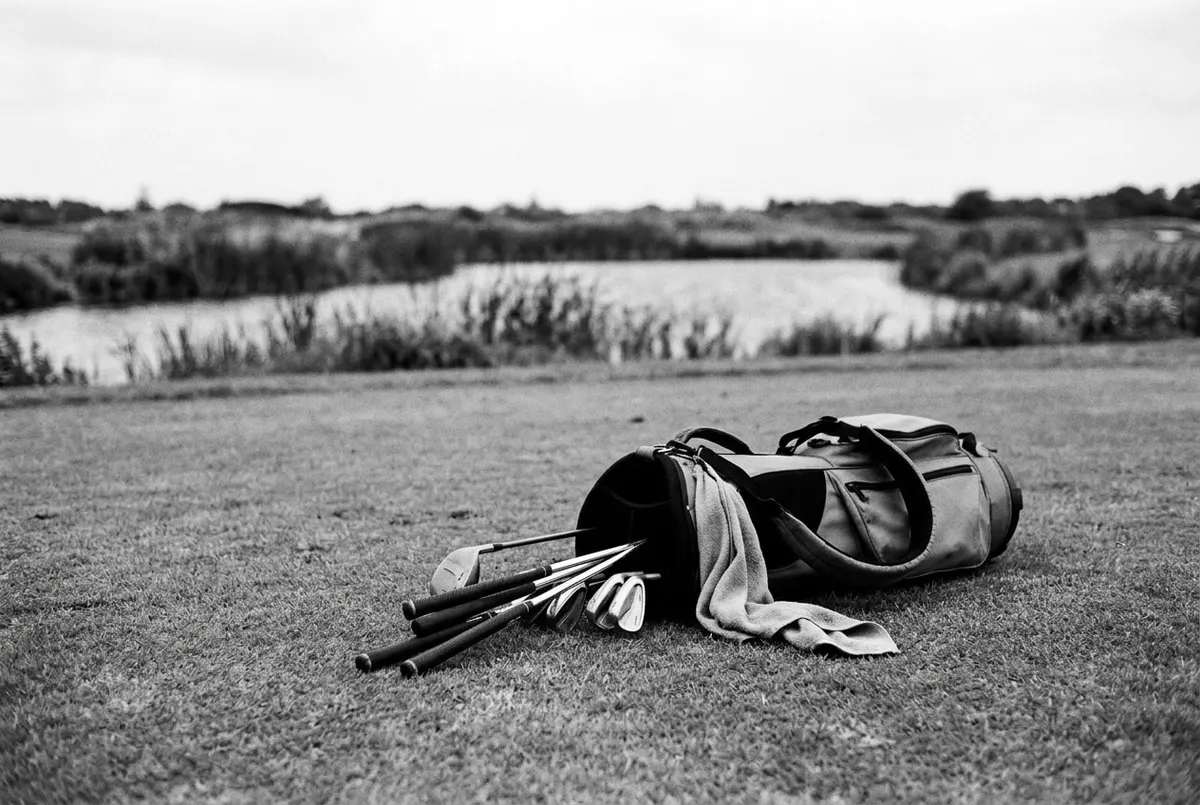 Een omgevallen golftas met clubs op de fairway, met een waterhazard op de achtergrond, in zwart-wit