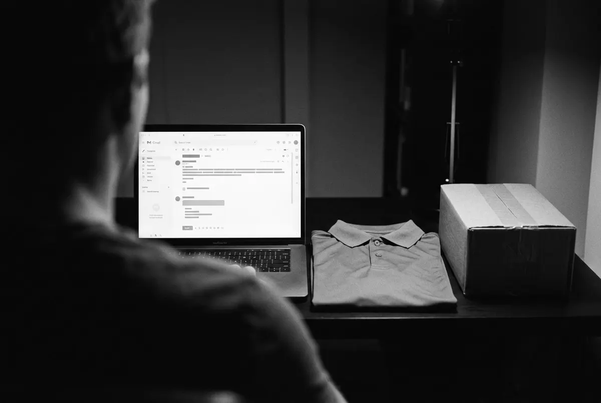 Laptop met een berichtenvenster op een bureau naast een opgevouwen golfshirt en een verzendpakket, in het donker.