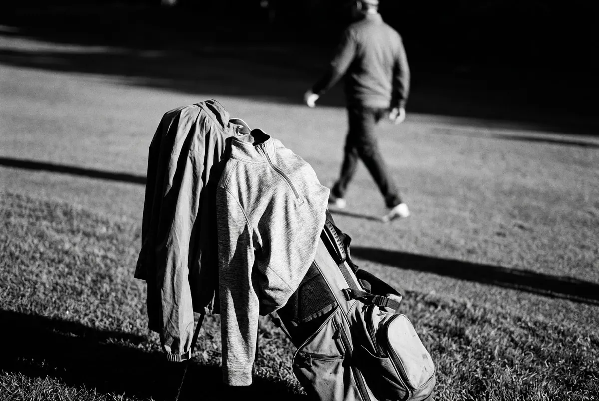 Regenjack en quarter-zip hangen over een golftas bij de eerste tee in herfstlicht