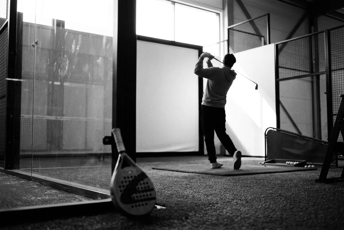 Een padelracket bij een padelbaan met op de achtergrond een golfer die afslaat op de fairway, in zwart-wit.