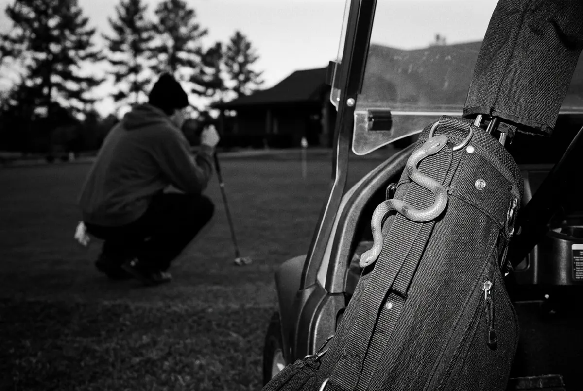 Zwart-witfoto van een rubberen speelgoedslang aan een golftas, met op de achtergrond een speler die een putt bekijkt.