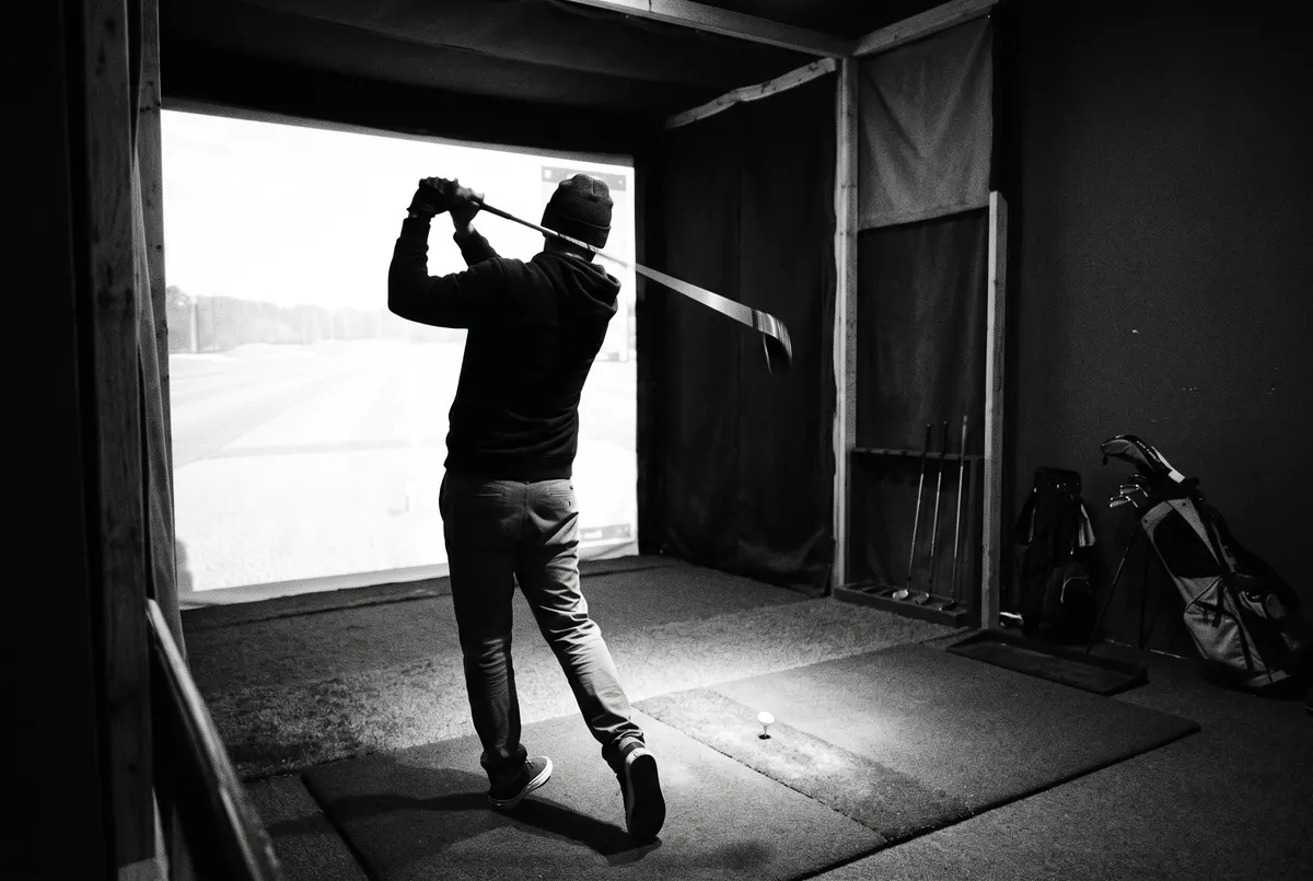 Silhouet van een golfer die een swing maakt in een donkere indoor simulatorbay, verlicht door het projectiescherm.