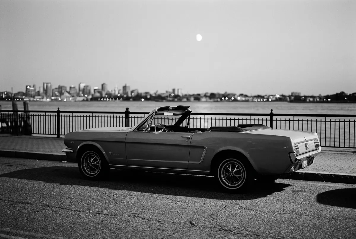 Een klassieke rode auto geparkeerd aan de kade van een haven, met een vage maan aan de hemel.