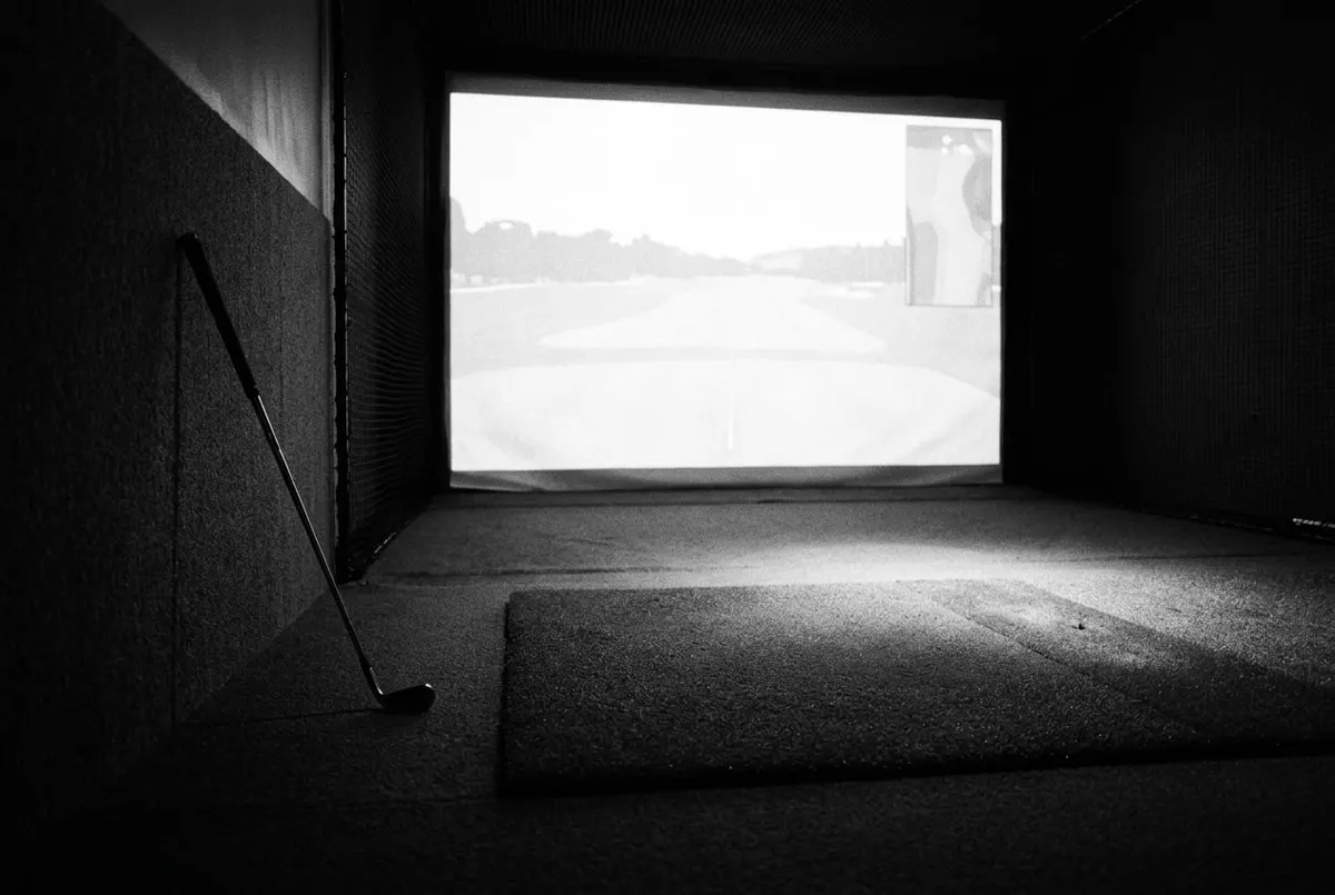 Lege golfsimulatorruimte met verlicht projectiescherm en een golfclub tegen de muur, in zwart-wit.