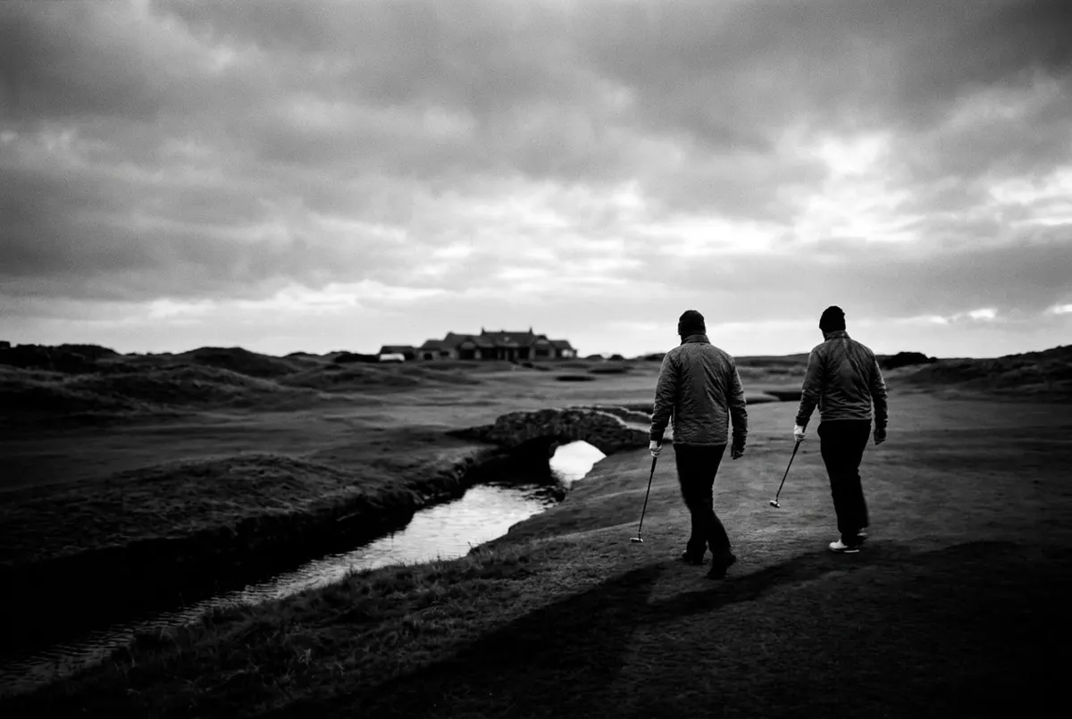 Twee golfers lopen bij avondlicht van de green af op een Schotse linksbaan, met op de achtergrond een stenen brug over een beekje.