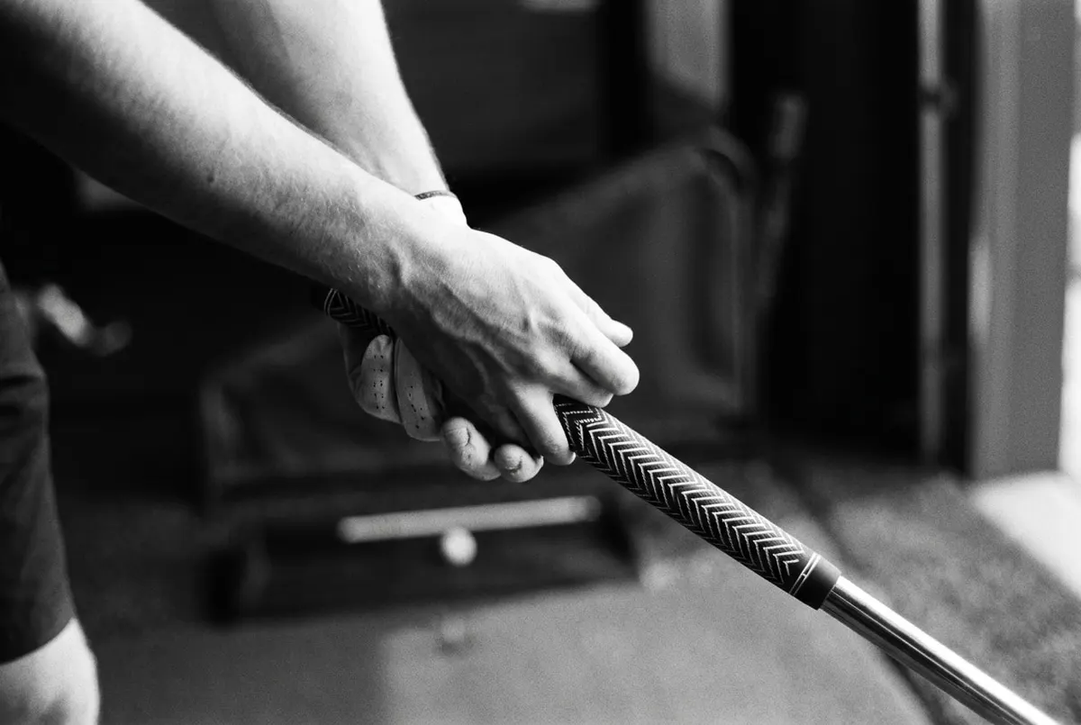 Close-up in zwart-wit van twee handen om een golfgrip met een opvallend patroon, de rest van de baan vervaagt op de achtergrond.