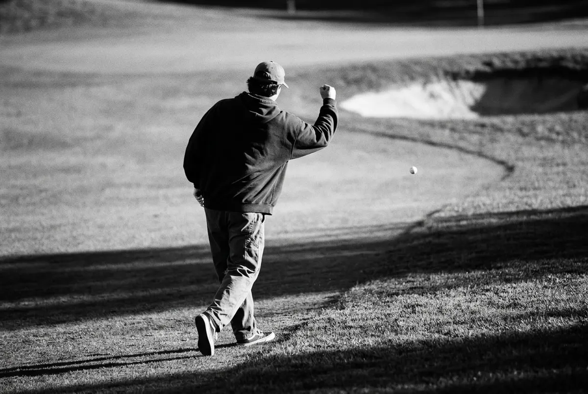 Golfer van achteren roept iets terwijl een bal nog in de lucht hangt richting een bunker