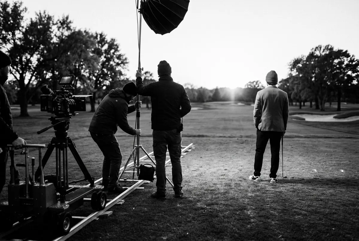 Filmcrew zet camera op bij een fairway tijdens gouden uur, golfer in streetwear staat met de rug naar de camera