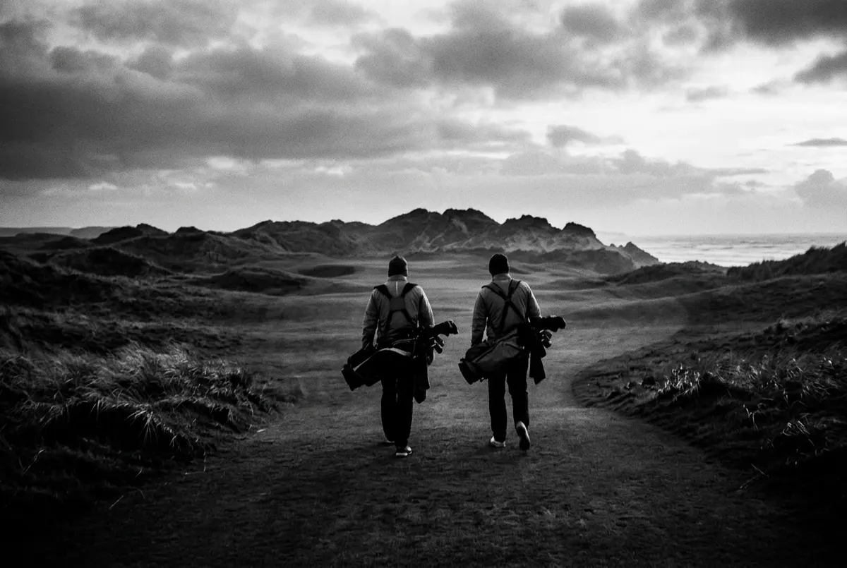 Twee golfers lopen over een mistige links golfbaan aan de Ierse kust, van achteren gefotografeerd.