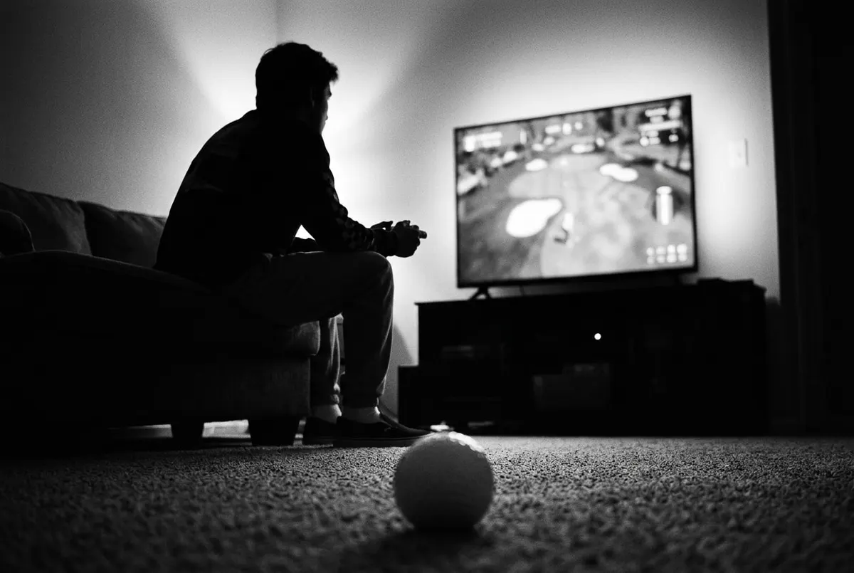 Silhouet van iemand met een controller op de bank, met een golfbal op de vloer op de voorgrond, in zwart-wit.