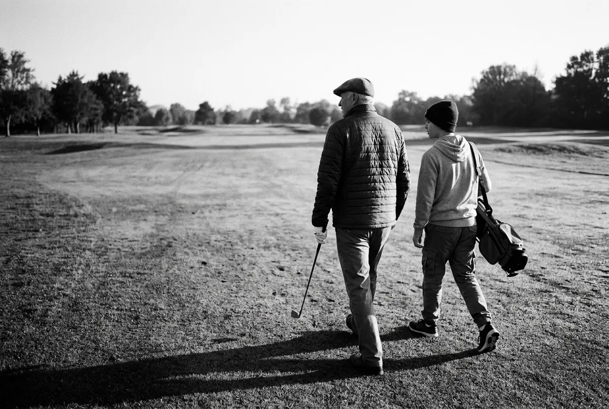 Zwart-witfoto van een opa en kleinzoon die samen over een golfbaan lopen, op de rug gezien, met een golftas over de schouder.