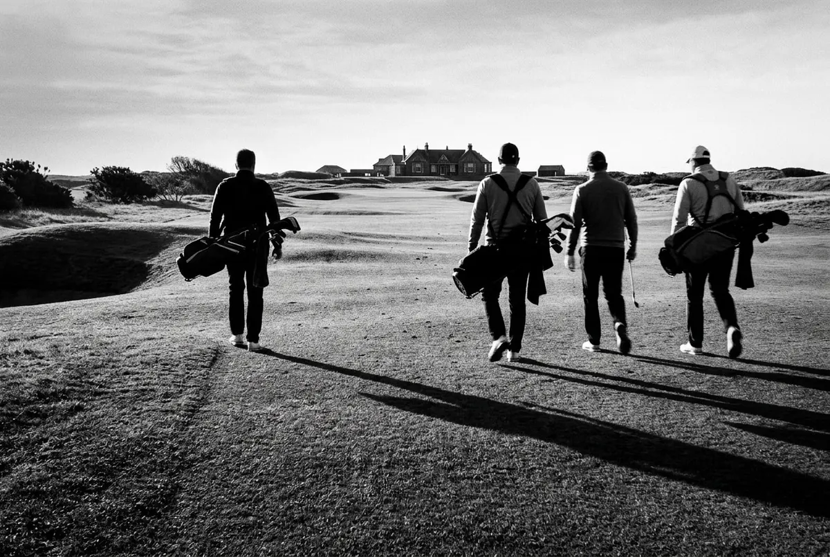 Vier anonieme golfers lopen over een Schotse links-golfbaan in ochtendlicht, van achteren gefotografeerd.