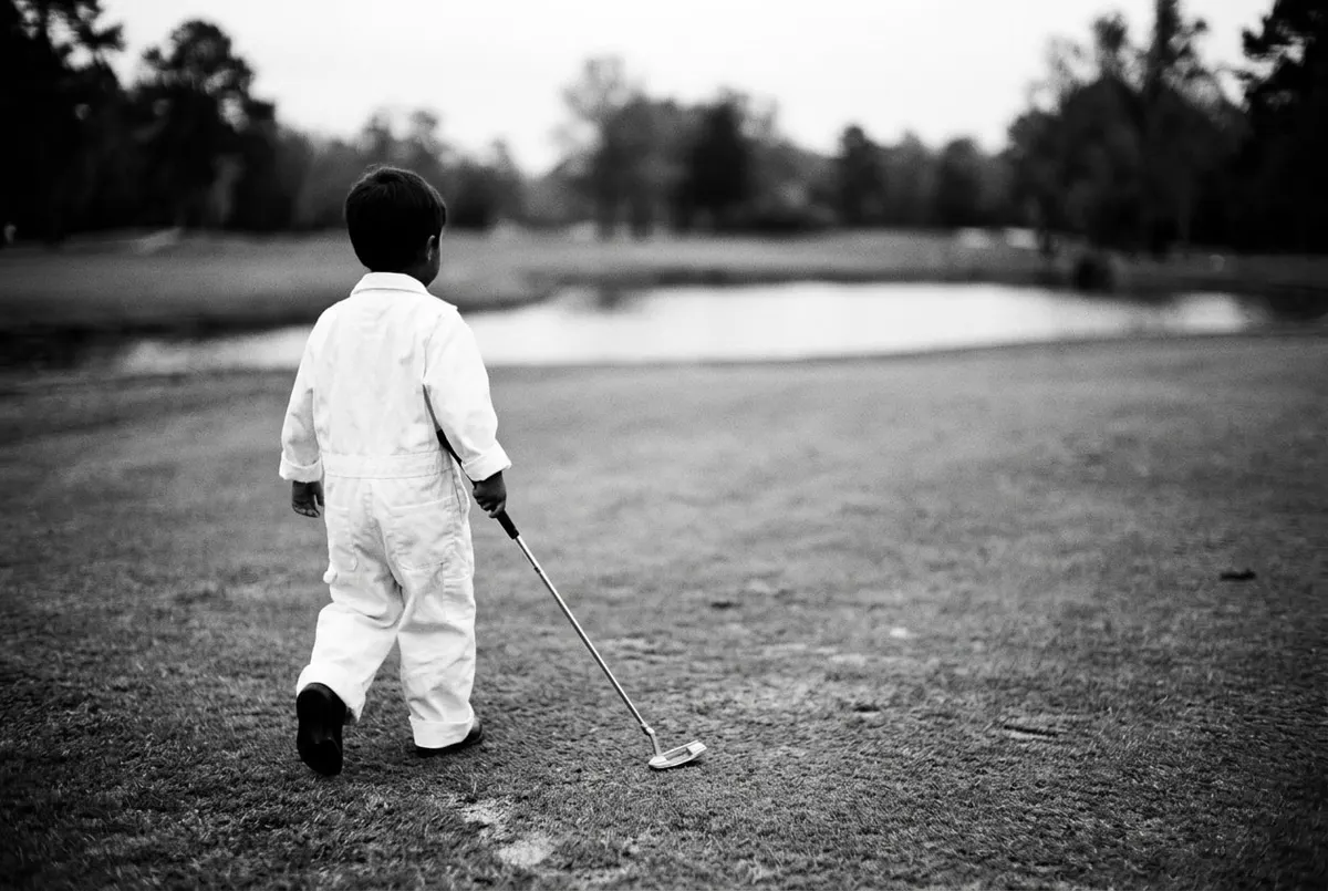 Kind in een witte caddiejumpsuit loopt met een putter over een golfbaan, van achteren gefotografeerd