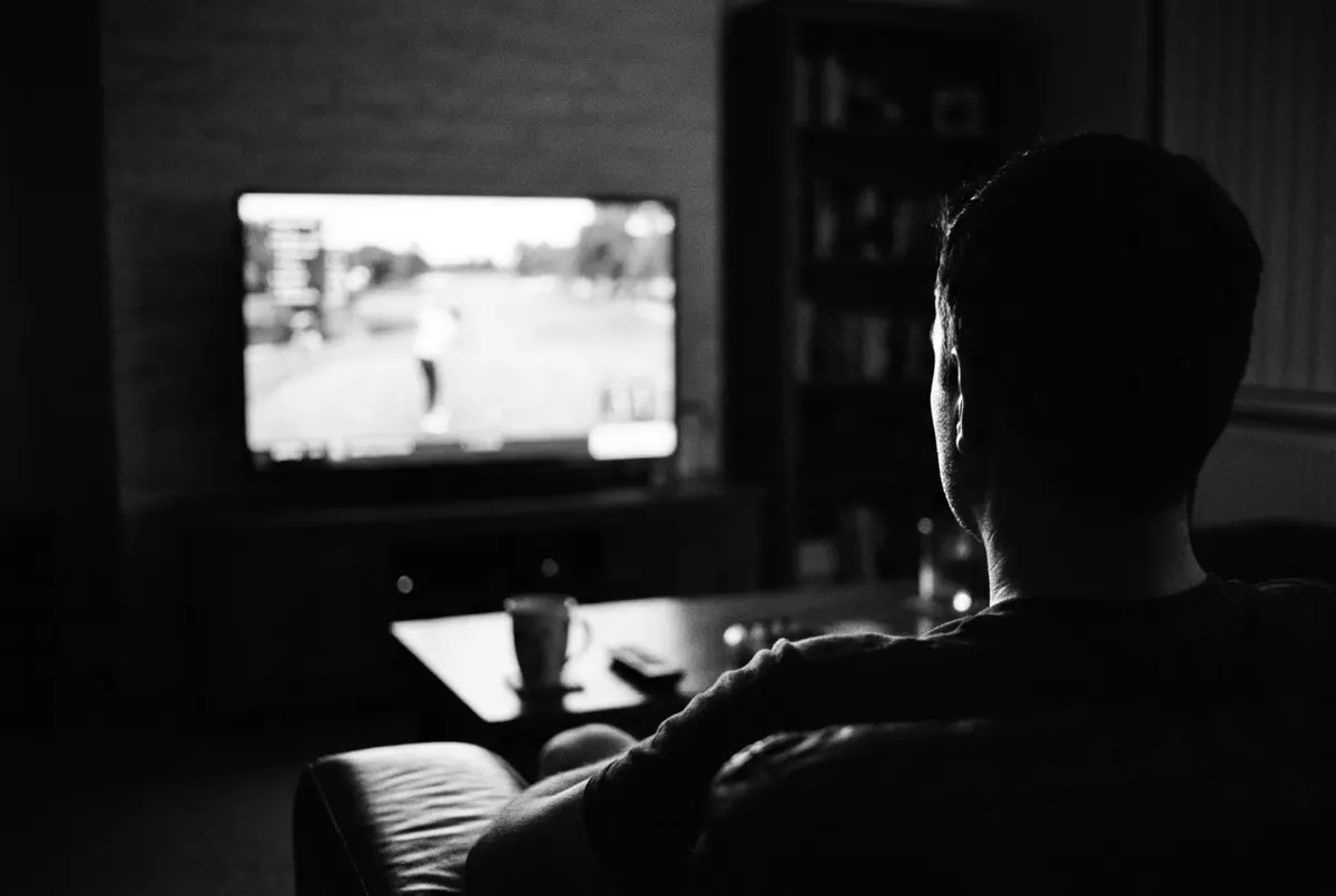 Zwart-witfoto van iemand die van achteren naar golfbeelden op tv kijkt in een donkere woonkamer.