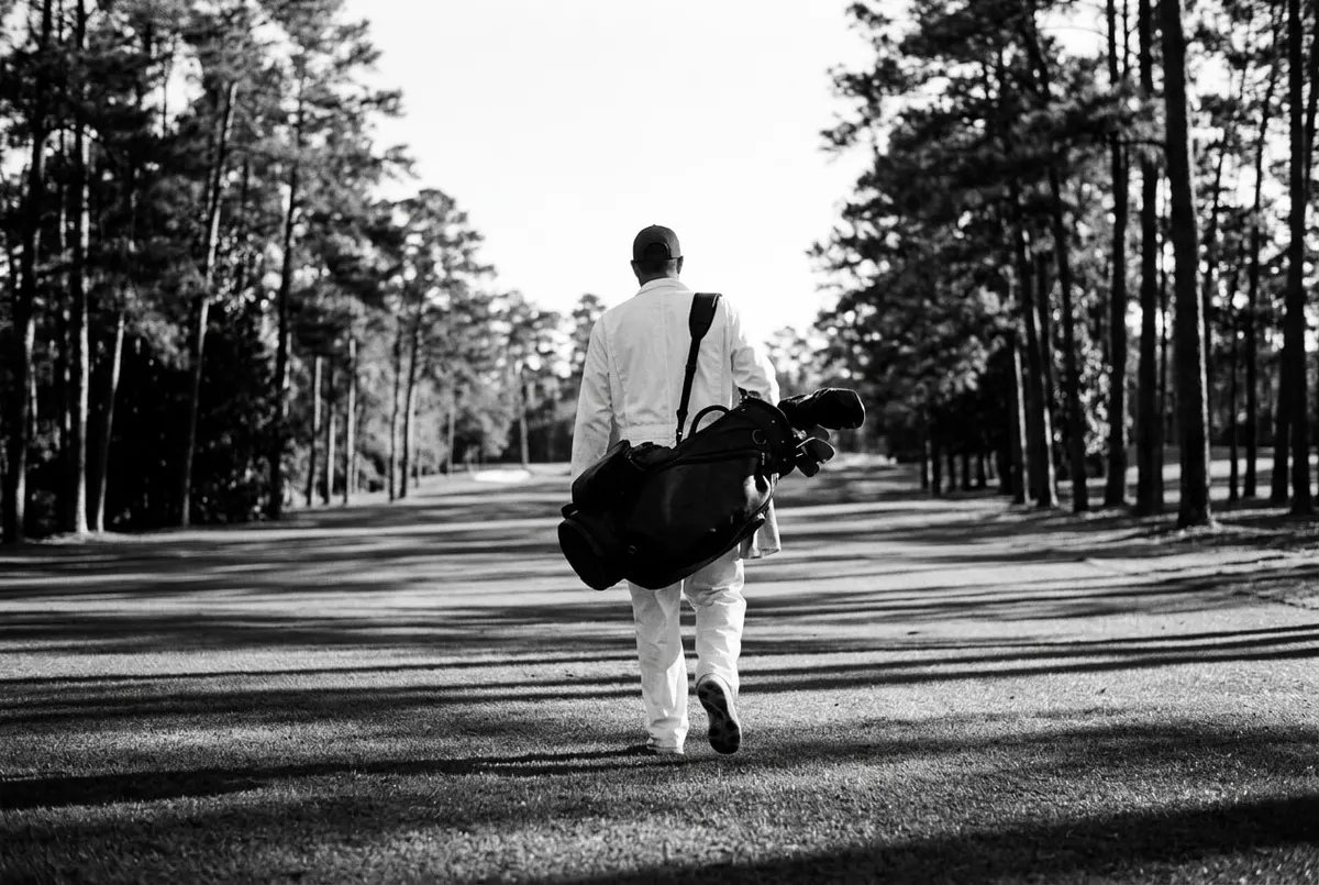 Caddie in witte overall loopt van achteren gefotografeerd met een golftas over een fairway tussen de bomen.