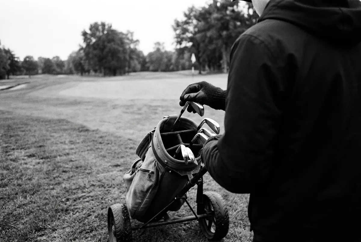 Zwart-witfoto van achteren van een caddie die golfclubs ordent in een tas op een trolley op een Nederlandse golfbaan.