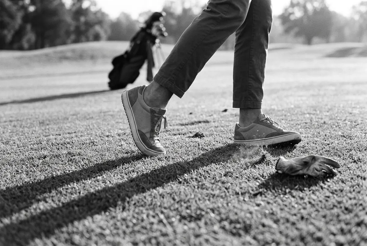 Close-up in zwart-wit van golfschoenen met een sneaker-silhouet op een fairway in ochtendlicht.